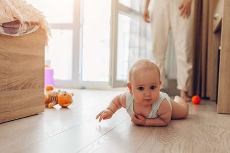 Infant crawling on floor in bedroom while mom watches. Little baby having fun at home. Kid looks at...