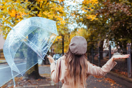Woman walking along autumn city street under transparent umbrella checking if it rains with hand. Fall season. Back viewの写真素材