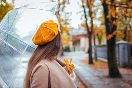 Stylish woman in fall clothes walking outdoors under transparent umbrella during rain. Autumn season weather. Back viewの写真素材