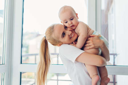 Young mother holding and hugging newborn baby son at home. Family relaxing on balcony. Infant wearing diaperの写真素材
