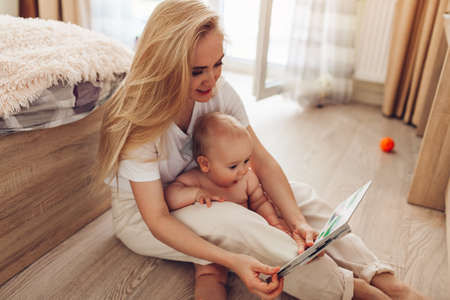 Woman reading to her baby son at home. Mother and kid sitting on floor with bookの写真素材