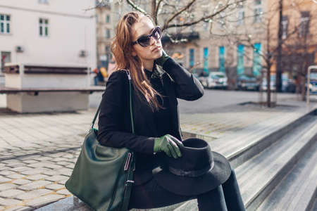 Stylish woman sitting on stairs in city. Girl wears hat, glasses with green handbag and gloves outdoors. Autumn fashion.の写真素材