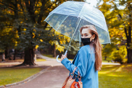 Stylish young woman in protective mask walking in autumn park under transparent umbrella after rain. Coronavirus covid-19 fashionの写真素材