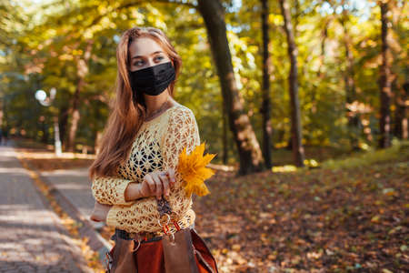 Pretty young woman wears black protective mask walking in autumn park holding bouquet of yellow leaves. Coronavirus covid-19 fashionの写真素材