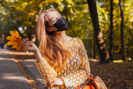Pretty young woman wears protective mask walking in autumn park holding yellow leaves. Coronavirus covid-19 fashion. Happy girl enjoys fall weatherの写真素材