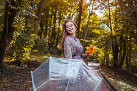 Happy young woman walking outdoors with transparent umbrella after rain holding yellow leaves. Fall season activities. Stylish girl enjoys weatherの写真素材