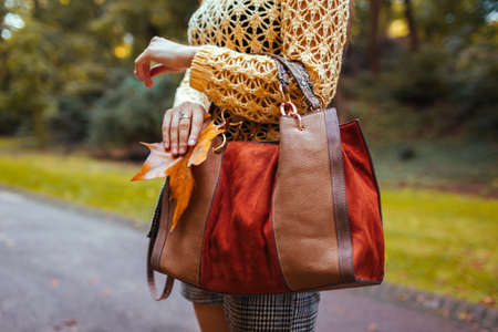 Close up of female leather handbag. Woman holds brown orange purse in park with yellow leaves....