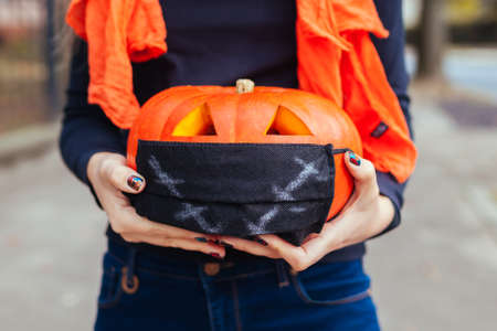 Halloween jack-o-lantern pumpkin in protective mask. Woman holds hand-made carved pumpkin outdoors. Celebration during coronavirus covid-19 pandemicの写真素材