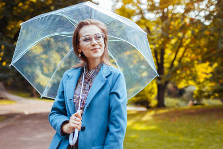 Stylish young woman in glasses walking in park with transparent umbrella wearing trendy outfit....