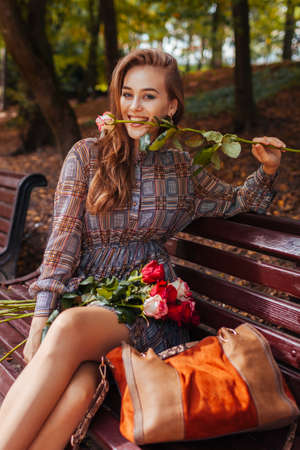 Beautiful woman sitting on bench with bouquet of roses in autumn park. Girl having fun biting rose flowerの写真素材