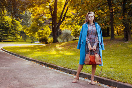 Full body portrait of stylish young woman walking in park with handbag wearing trendy outfit. Autumn...