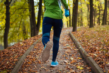 Runner working out in autumn park wearing sportive clothes. Young woman running with water bottle among yellow trees. Close up of shoesの写真素材