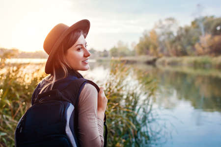 Traveler with backpack relaxing by autumn river at sunset. Happy young woman enjoys fall weather and landscape. Active lifestyleの写真素材