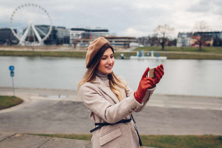 Stylish woman taking selfie on phone walking along pier by Wisla river in Krakow, Poland enjoying...