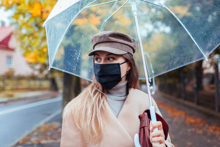 Young woman in protective mask walking along autumn city street under transparent umbrella during rain. Covidの写真素材