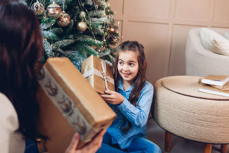 Merry Christmas and Happy New Year. Mom and daughter shaking gift boxes under tree at home. Excited familyの写真素材