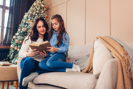 Family spending time at home for Christmas and New year holidays. Mother and daughter reading book by Christmas treeの写真素材