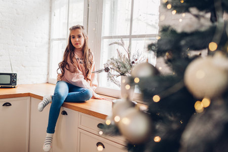 Little girl sitting on window sill by Christmas tree on kitchen. Stay at home for New Year winter holidaysの写真素材