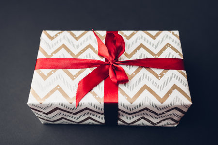 Christmas and New year gift box wrapped in festive paper and decorated with red ribbon with bow on black background. Winter holidays presentの写真素材