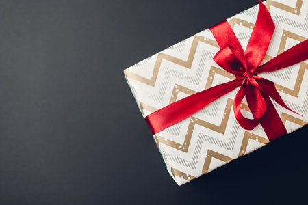 Christmas and New year gift box wrapped in holiday paper and decorated with red ribbon with bow on black background. Spaceの写真素材
