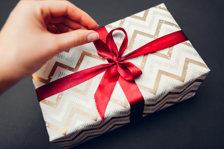 Woman opens Christmas and New year gift box wrapped in paper and decorated with red ribbon bow on black background. WInter holiday presentの写真素材