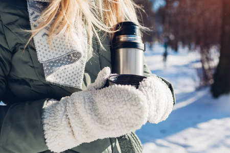 Woman drinking hot tea holding vacuum flask in winter park. Drinks to warm up in snowy frosty weather outdoorsの写真素材