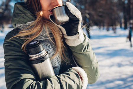 Woman drinking hot tea holding vacuum flask in winter park. Drinks to warm up in snowy frosty weather outdoorsの写真素材