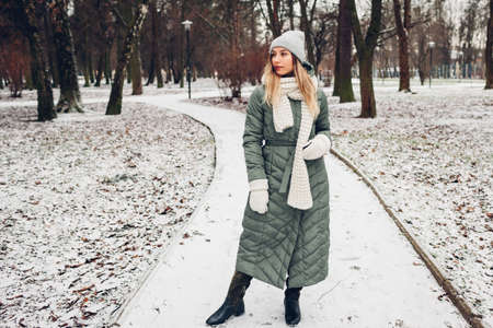 Winter fashion. Young woman wearing long green coat with scarf, hat, mittens and boots in snowy park. Modern clothesの写真素材
