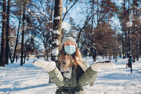 Winter portrait of happy woman in protective mask playing with snow outdoors. virus  pandemic new reality. Girl having fun throwing snowの写真素材