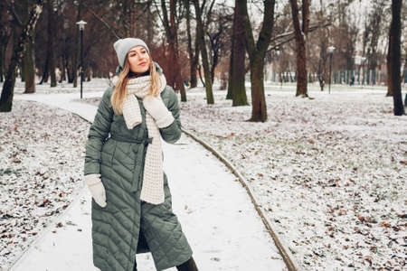 Winter fashion. Portrait of young woman wearing long green coat with scarf, hat, mittens in snowy park. Modern clothesの写真素材