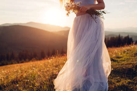Bride wearing blue wedding dress holding bouquet in mountains at sunset. Woman walking on meadow in chamomile flowersの写真素材