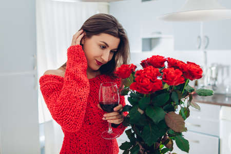 Young woman found bouquet of red roses flowers on kitchen from boyfriend. Happy girl drinking wine at home. Valentine's day surpriseの写真素材