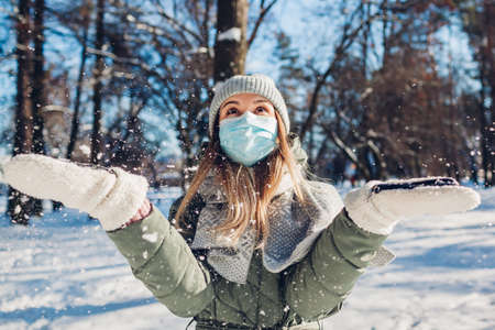 Winter portrait of happy woman in protective mask playing with snow outdoors.