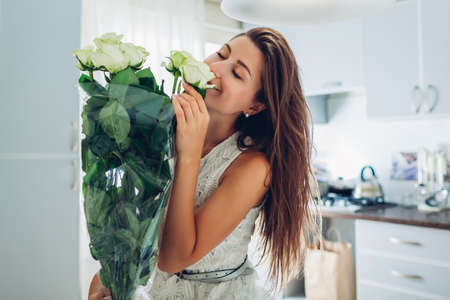 Valentine's day present. Happy young woman smelling bouquet of roses flowers from husband on kitchen. Women's day. Surprise from boyfriendの写真素材