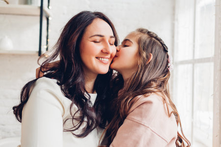 Mother and daughter hugging on kitchen window sill. Girl kissing mom on cheek. Family having good time together at home. Mother's dayの写真素材