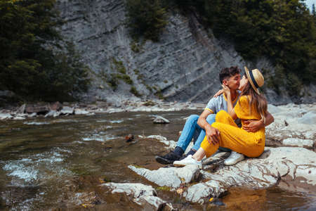 Couple of tourists kissing by mountain river enjoying landscape. Travelers hugging sitting on rock. Summer vacation trip. Back viewの写真素材