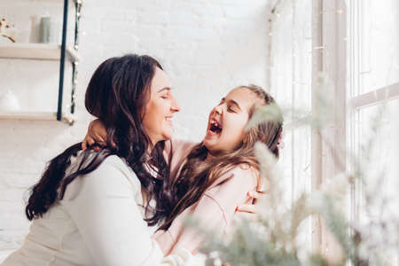Mothers day. Mother and daughter laughing and hugging on kitchen window sill. Family having fun together at homeの写真素材
