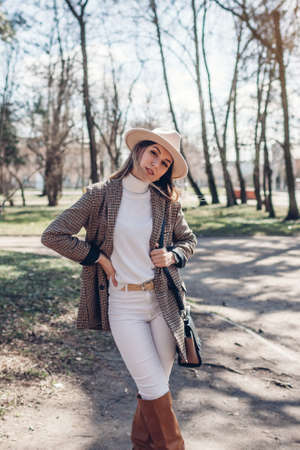 Beautiful woman wearing stylish blazer, hat and boots holding purse in park. Spring female fashionable outfit.の写真素材