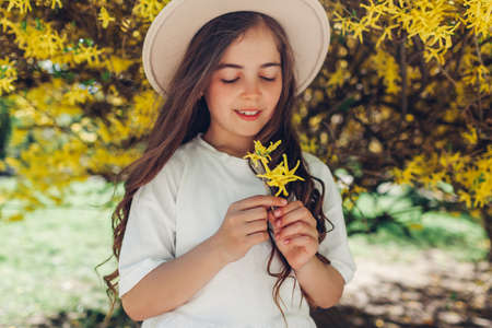 Stylish little smiling girl holding yellow flowers under blooming forsythia bush in spring garden. Kid wearing hatの写真素材