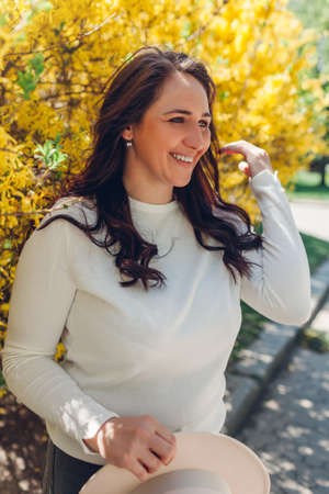 Portrait of smiling middle-aged woman walking in blooming spring garden. Happy stylish lady holding hat outdoors surrounded with flowersの写真素材