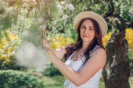 Portrait of middle-aged woman walking in blooming spring garden. Happy stylish lady wearing hat and white dress outdoors surrounded with flowersの写真素材