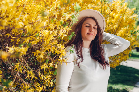 Portrait of smiling middle-aged woman walking in blooming spring garden. Happy stylish lady smelling...