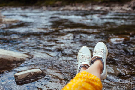 Woman relaxes by mountain river enjoying landscape. Traveler sitting on rock. Summer vacation. Close up of shoesの写真素材
