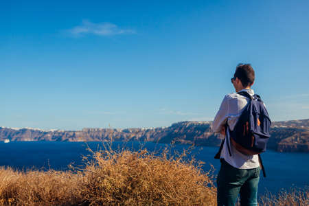 Man traveler admires seaside landscape on Santorini island. Tourist backpacker enjoys Caldera view and mountainsの写真素材