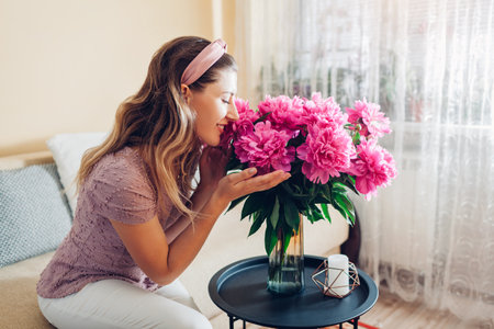 Woman smelling peonies flowers in vase at home. Housewife enjoys bouquet of fresh pink blooms. Interior and decor of living roomの写真素材