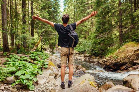 Traveler hiker with backpack raising arms by mountain river in Carpathian forest feeling happy. Man tourist enjoys landscape. Trip to summer Ukraineの写真素材