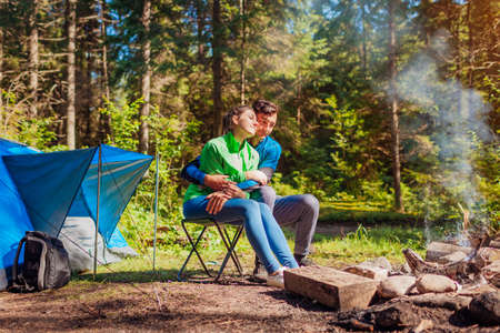Young couple of travelers enjoy campfire in summer forest. Tourists sitting by tent hugging. Camping in woodsの写真素材