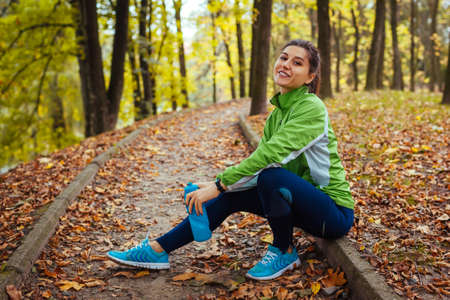 Runner having rest after workout in autumn park. Happy woman holding water bottle sitting on path. Sportive active healthy lifestyle. Fall workout on fresh airの写真素材