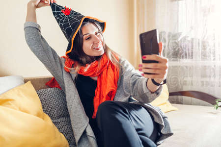 Halloween. Woman in witch hat taking selfie using smartphone. Celebrating holidays at home during lockdownの写真素材