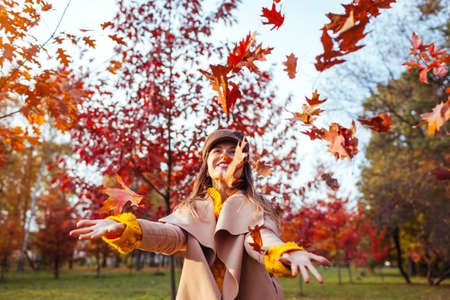 Fall season activities. Woman throwing leaves in autumn park. Young woman having fun among red trees...